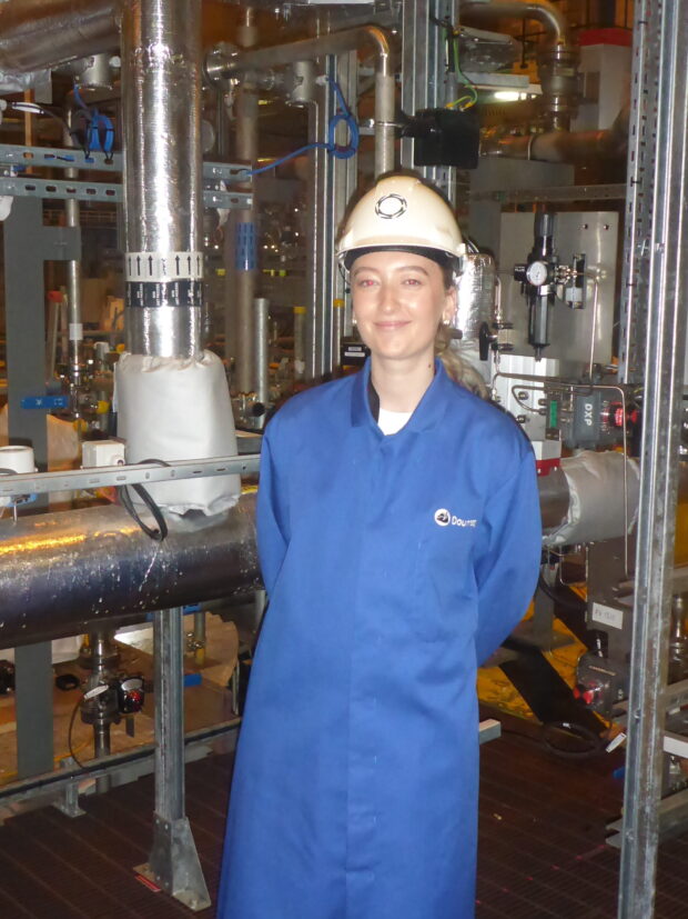 Woman stood with Dounreay branded overalls and hard hat on site with pipes in the background