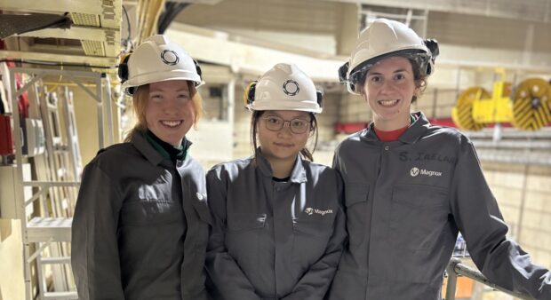 Three graduates stood inside the Dragon Reactor hall wearing hard hats and PPE.